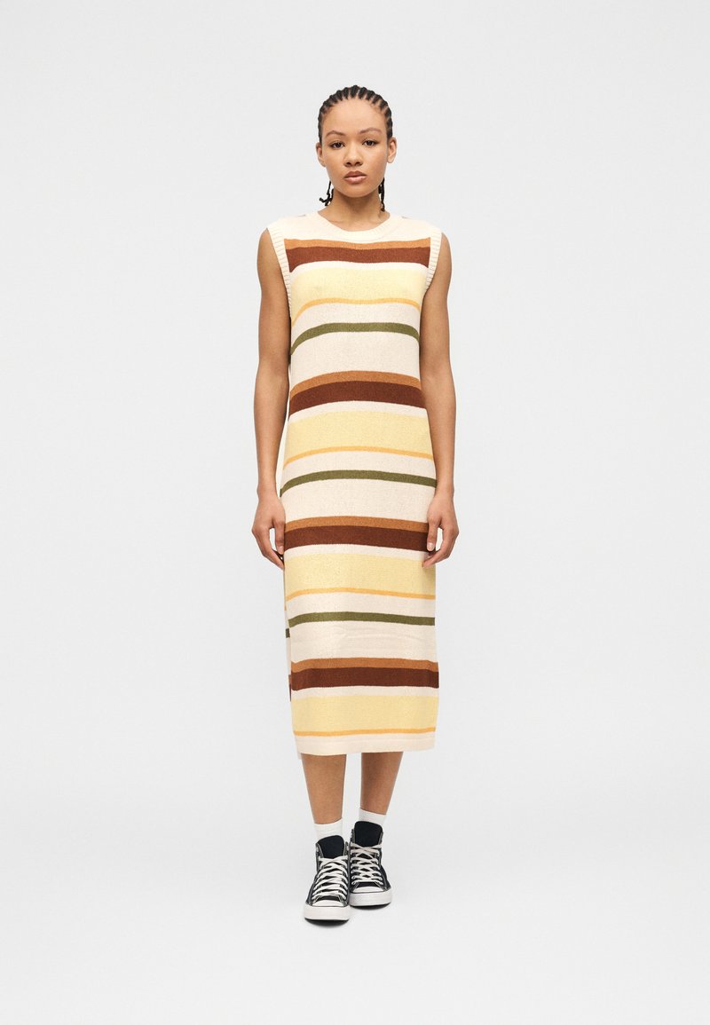 Γυναίκα με πλεξούδες που φοράει αμάνικο ριγέ πλεκτό φόρεμα σε καφέ, κίτρινους, κρεμ και ελιά αποχρώσεις, σε συνδυασμό με μαύρα ψηλά αθλητικά παπούτσια.