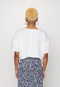 Camisa branca, curta, de mangas curtas, com fecho de botão nas costas. Combinada com uma saia azul estampada com um design abstrato.