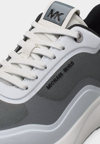 Κοντινό πλάνο λευκού και γκρι αθλητικού παπουτσιού Michael Kors με λευκά κορδόνια, δίχτυα και το όνομα της μάρκας στο πλάι και στη γλώσσα του παπουτσιού.