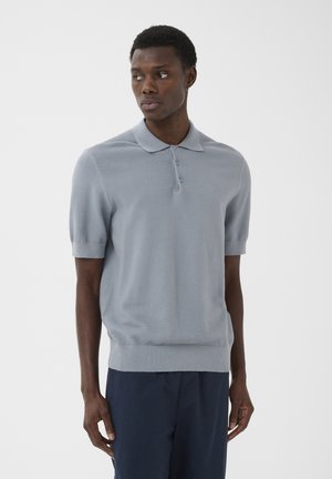 Junger Mann, der ein hellgraues kurzärmeliges Polohemd und dunkelblaue Hosen trägt, steht vor einem schlichten weißen Hintergrund und schaut nach links.