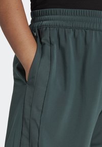 Sportovní kalhoty z tmav�ého modrozeleného materiálu s elastickým pasem, boční kapsou a hladkou texturou, s jemným skladem podél strany.