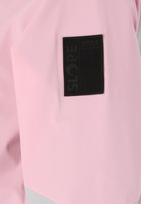 Rosa vattentät jacka med en slät textur, försedd med en svart rektangulär märke som är märkt med "SLOPE STYLE" på vänster ärm.