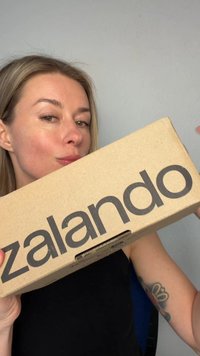 Vrouw met licht haar houdt een Zalando kartonnen doos vlak bij haar gezicht en toont een tatoeage op haar bovenarm tegen een effen achtergrond.
