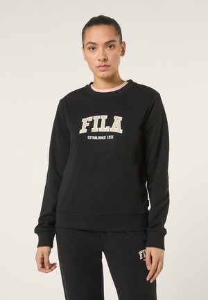 Vrouw draagt een zwarte FILA-trui en bijpassende zwarte broek, staand tegen een effen lichte achtergrond, naar voren gericht.