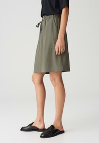 Woman wearing olive green knee-length skirt with drawstring waist and black slip-on loafers, standing against plain background.