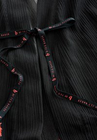 Zwarte geplooide wikkeljurk met een gestructureerd oppervlak en een contrasterende lintstrik met "b by Ted Baker" in rode letters.