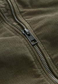 Primer plano de un zipper metálico parcialmente desabrochado sobre una tela de pana verde oliva.