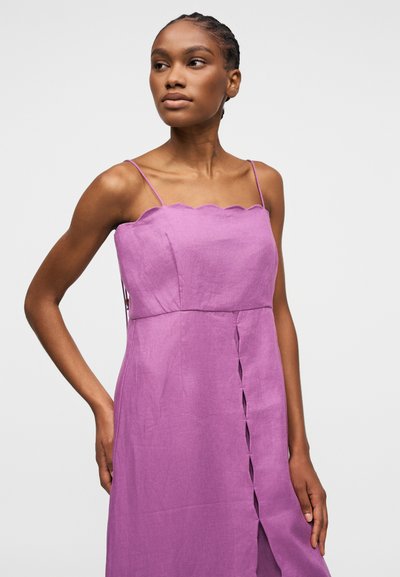 Jeune femme aux cheveux tressés portant une robe violette sans manches avec de fines bretelles et un décolleté à bords festonnés, debout devant un fond clair uni.