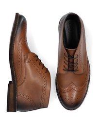 Brązowe skórzane buty z designem typu wingtip, detalami brogu oraz z warstwowym obcasem. Tekstura jest gładka z subtelnymi gradientami.