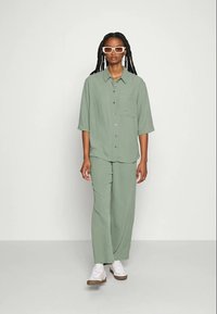 Απαλή πράσινη, χαλαρή μπλούζα με τσέπη μπροστά και κοντά μανίκια, συνδυασμένη με αντίστοιχες φαρδιές παντελόνες. Φοράει λευκά παπούτσια και γυαλιά.