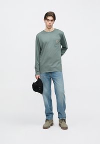 Πράσινο βαμβακερό πουκάμισο με μακριά μανίκια και τσέπη, ανοιχτό μπλε τζιν, γκρι μπότες και μαύρο καπέλο. Διαθέτει λογότυπο στην τσέπη του πουκάμισου.