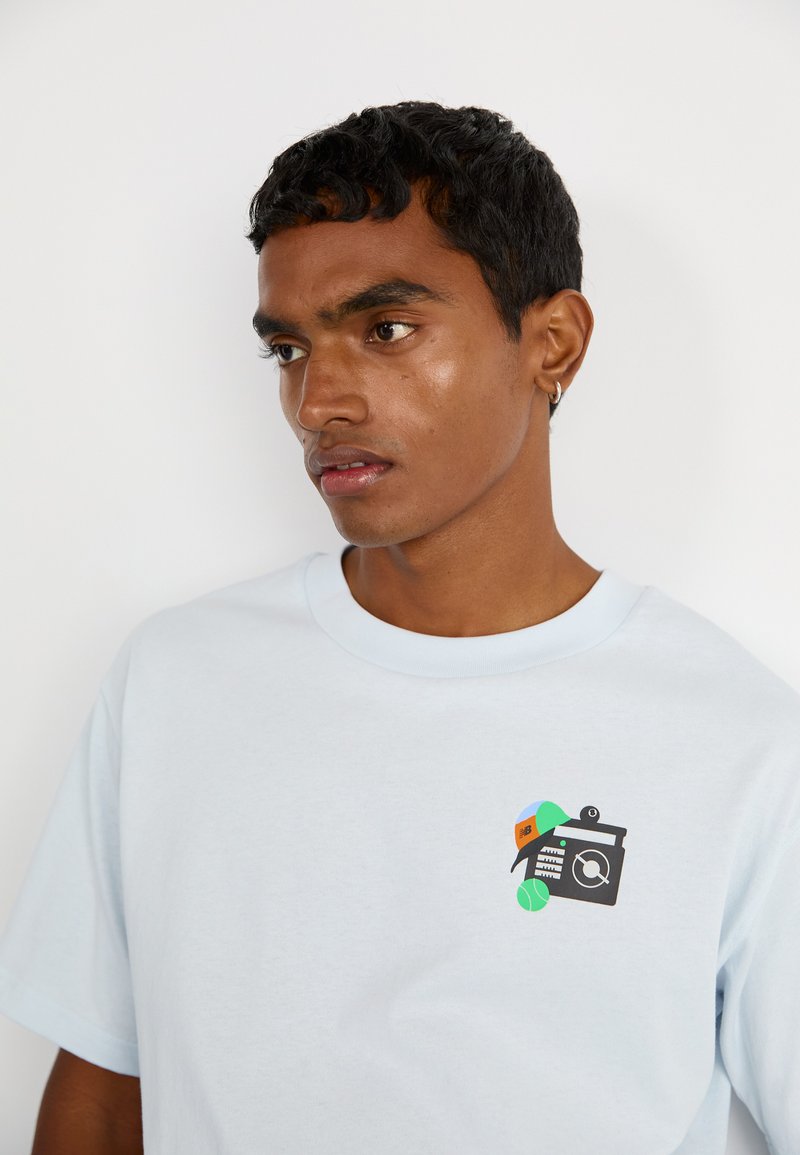 Camiseta de algodón azul claro con cuello redondo, que presenta una impresión gráfica colorida de una cámara, un portapapeles y una pelota de tenis en la parte frontal.