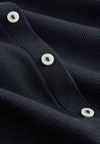 Tengerészkék bordázott anyag fehér gombokkal, melyeken kis fekete pötty található. A szövet sima textúrájú és enyhén fényes, jellegzetes vízszintes vonalakkal.