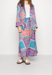 Barevný vzorovaný blazer a sukně v modrých, oranžových a růžových odstínech s volným střihem, doplněné o bílé skluzné sandály.