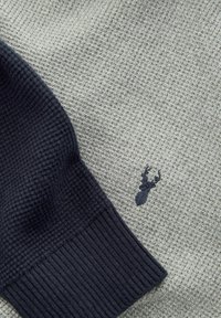 Strikket sweater med en struktureret lys grå krop og mørkegrå ribbede mansjetter, med et lille broderet hjorte-logo på brystet.