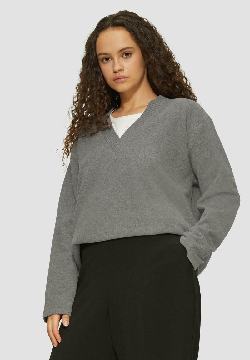 Grauer V-Ausschnitt-Pullover aus leichtem Baumwollmaterial mit langen Ärmeln und einer lockeren Passform, kombiniert mit hochtaillierten schwarzen Hosen.