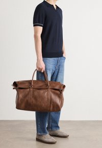 Sac de voyage en cuir marron à texture lisse, tenu par une personne. Il possède deux poignées robustes et une fermeture supérieure zippée.