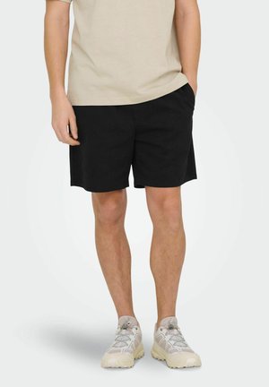 Mężczyzna w beżowej koszulce, czarnych szortach z bocznymi kieszeniami i jasnych sznurowanych trampkach, stojący z jedną ręką w kieszeni.