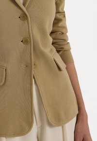 Beige bleiseri teksturoituu neulottua kangasta notched-kauluksella, kolmella painikkeella ja sivutaskuilla. Ominaisuuksiin kuuluu muotoutunut istuvuus ja puhtaat viivat.