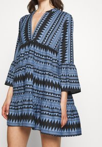 Robe à motifs géométriques bleu et noir avec un décolleté en V profond, des manches évasées et une jupe évasée, réalisée dans un tissu léger.