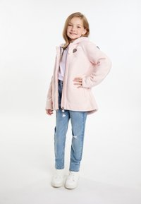 Giacca in pile rosa chiaro con cappuccio, chiusura a zip, due tasche laterali e accessori decorativi; abbinata a jeans blu strappati e sneakers bianche.