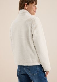 Femme de profil portant une veste en peluche blanche à col haut et un jean bleu sur un fond neutre.