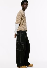 Un jeune homme se tient de profil, portant une chemise beige à rayures à manches courtes sur un haut à manches longues blanc, un pantalon cargo noir et des bottes beiges.
