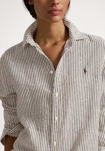 Sieviete, tērpusies bēša un baltā vertikāli svītrotā kreklā ar apkakli un uzruļētām piedurknēm.