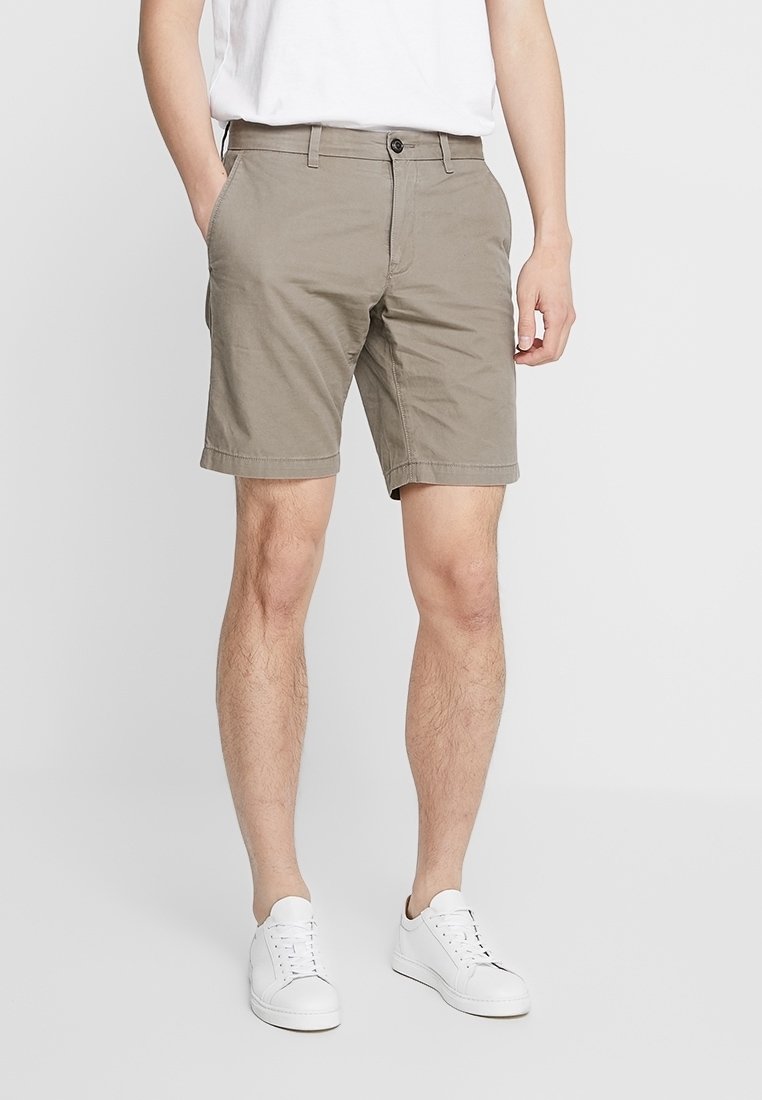 Shorts en coton beige à coupe droite, taille plate, avec deux poches latérales. Portés avec un t-shirt blanc et des baskets blanches.