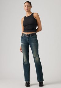 Débardeur noir côtelé assorti à un jean bleu foncé avec des détails usés et une ceinture noire. Le modèle porte des bottines noires.