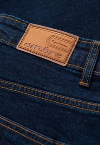 Jeans en denim bleu foncé avec une étiquette en cuir crème embossée des mots "ombre" et "Denim Premium", avec des coutures contrastantes orange.