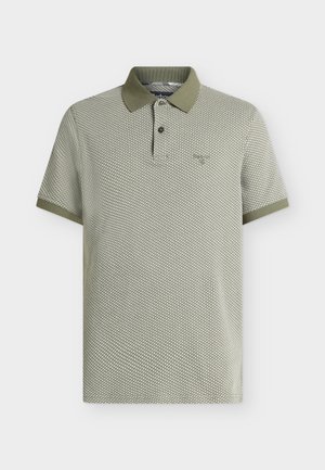 Jeune homme aux cheveux bouclés, debout les mains dans les poches, portant un polo gris à motifs, un pantalon foncé et ample, une ceinture marron et des chaussures foncées.