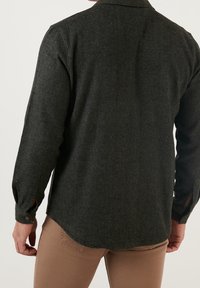 Camisa verde oscuro hecha de un tejido texturizado, con cuello, mangas largas y puños abotonados, y con un dobladillo ligeramente curvado.