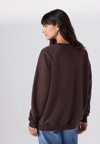 Brūns pārmērīgs džemperis ar apaļu kakla izgriezumu, karenām plecu šuvēm un ribotām aprocēm, kombinēts ar zilu džinsu fona neitrālā vidē.