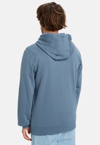 Světle modrá mikina s velkou kapucí, dlouhými rukávy a volnějším střihem. Měkká textura látky, s rovnými lemy a božním štítkem.