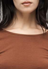 Κοντινό πλάνο του κάτω μέρους προσώπου γυναίκας, του λαιμού και των ώμων με καφέ τοπ χωρίς ώμους, τα μαύρα μαλλιά πλαισιώνουν τις πλευρές.