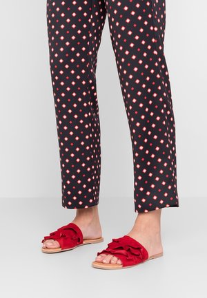 Piernas inferiores con pantalones negros con patrones geométricos en rojo y blanco, y sandalias rojas con volantes sin talón sobre un fondo claro.