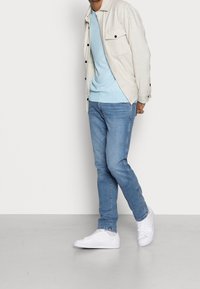 Jasnoszara koszula na guziki nałożona na jasnoniebieski sweter, do tego niebieskie dżinsy i białe sneakersy. Gładkie tekstury i swobodny design.