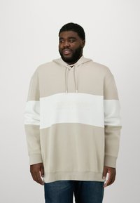Beige und weiße Kapuzenjacke mit strukturierten Streifen und einer Vordertasche. Aus weichem Stoff gefertigt, mit Kordelzug und Logo. Kombinierbar mit blauen Jeans.