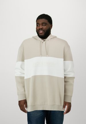 Beige und weiße Kapuzenjacke mit strukturierten Streifen und einer Vordertasche. Aus weichem Stoff gefertigt, mit Kordelzug und Logo. Kombinierbar mit blauen Jeans.