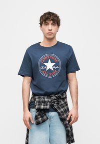 Νεαρός άνδρας που φοράει μπλε μπλουζάκι Converse All Star, ανοιχτόχρωμο μπλε τζιν και πουκάμισο μαύρο με γκρι καρό δεμένο γύρω από τη μέση του.