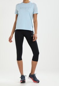 Personne portant un t-shirt Nike bleu clair, un legging cropped noir avec des logos Nike, et des chaussures de course noires avec des semelles rouges et blanches.