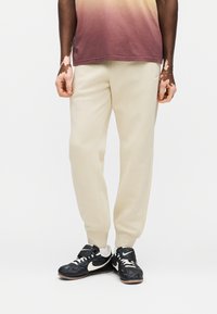 Φόρμες εκρού με ριμπ μανσέτες και λογότυπο Nike. Φορεμένες με μαύρα αθλητικά παπούτσια με λευκές λεπτομέρειες και λουράκι με Velcro.