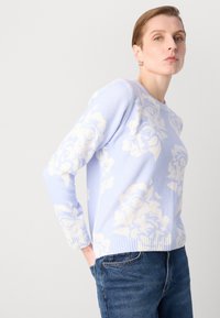Lys blå genser med hvitt blomsterdesign, laget av mykt materiale. Har rund hals og ribbede mansjetter, kombinert med mørk denim.