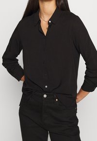 Chemise noire à boutons avec col, manches longues et coupe décontractée. Présente une texture lisse et des boutons contrastés. Associée à un jean noir.