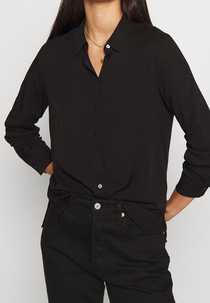 Chemise noire à boutons avec col, manches longues et coupe décontractée. Présente une texture lisse et des boutons contrastés. Associée à un jean noir.