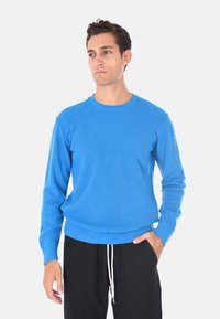 Maglione a coste blu con maniche lunghe e scollatura rotonda, con una finitura testurizzata. Indossato sopra pantaloni neri con coulisse.