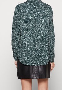 Camicia a maniche lunghe di colore verde scuro con pois bianchi, dotata di colletto classico e orlo dritto, indossata sopra una gonna di pelle nera.