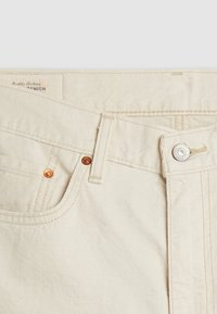 Gros plan sur un jean en denim beige clair montrant la poche avant, la boucle de ceinture, le bouton en métal et les rivets avec la texture visible du tissu et les coutures.