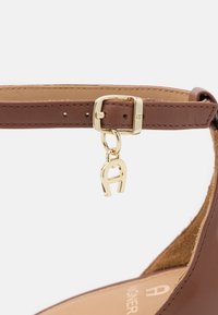 Bride de cheville en cuir marron avec une boucle en métal doré et un petit charme doré en forme de fer à cheval suspendu à la boucle.